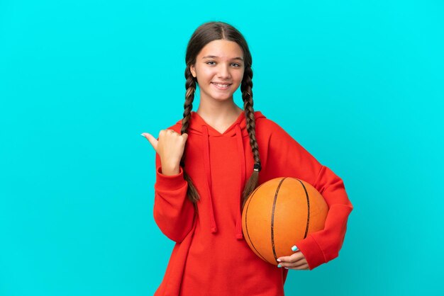 Niña caucásica jugando baloncesto aislada de fondo azul apuntando hacia un lado para presentar un producto