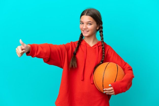 Niña caucásica jugando baloncesto aislada de fondo azul dando un gesto de pulgar hacia arriba