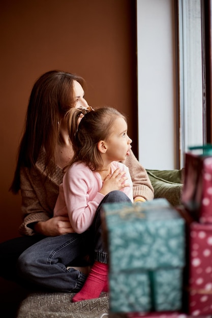 Niña caucásica con madre mirando la ventana y soñando Muchas cajas de regalo alrededor del concepto de Navidad familiar feliz