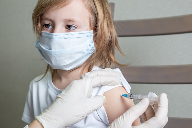 Niña caucásica con una máscara médica se sienta en una silla en la cabina del médico y recibe una vacuna, mira al médico