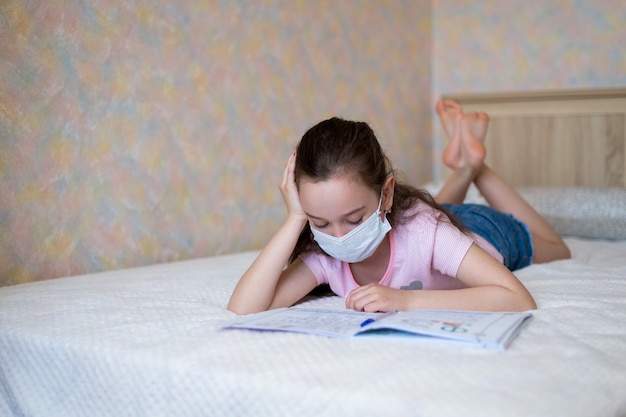 Niña caucásica en una máscara protectora escribe en un cuaderno durante la cuarentena