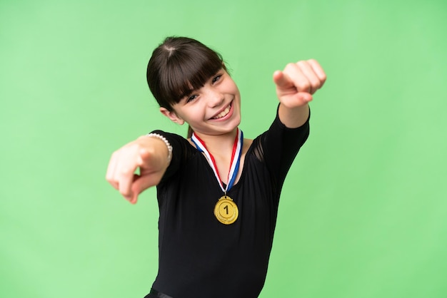 Foto una niña caucásica con medallas sobre un fondo aislado te señala con el dedo mientras sonríe
