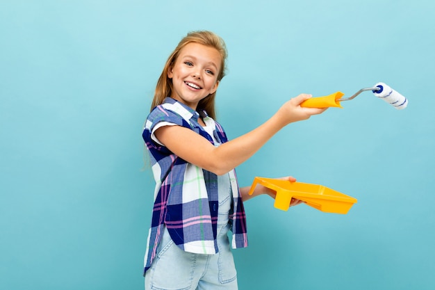 Niña caucásica pinta una pared con un rodillo y pintura aislado sobre fondo azul.