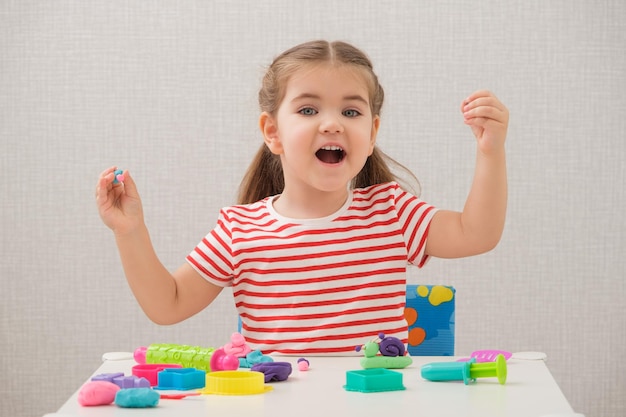 Niña caucásica rubia con camiseta brillante a rayas está jugando con plastilina en casa modelando arcilla