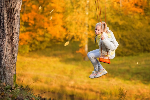 Niña caucásica rubia feliz sonriendo y montando un columpio de cuerda en otoño en el parque