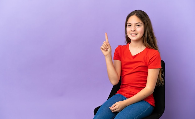 Niña caucásica sentada en una silla aislada sobre fondo púrpura apuntando hacia una gran idea