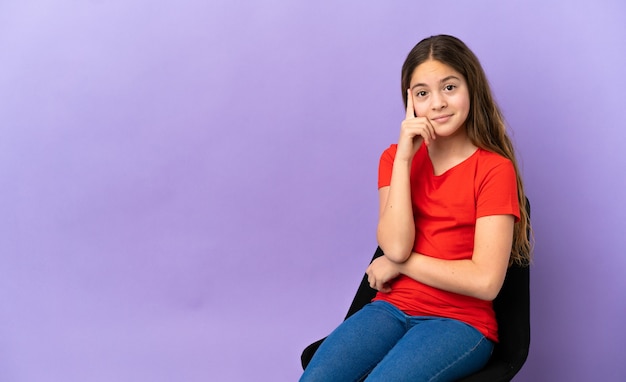 Foto niña caucásica sentada en una silla aislada sobre fondo púrpura pensando en una idea