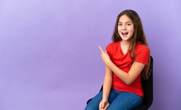 Niña caucásica sentada en una silla aislada sobre fondo púrpura sorprendido y apuntando hacia el lado