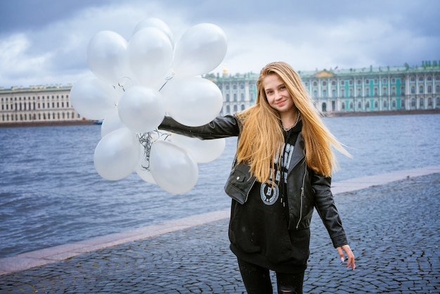 Niña caucásica sosteniendo globos blancos de pie junto al río en el terraplén de la ciudad en una nube ...