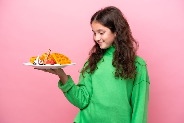 Niña caucásica sosteniendo gofres aislados en fondo rosa con expresión feliz