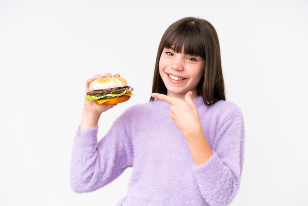 Niña caucásica sosteniendo una hamburguesa sobre un fondo aislado y señalándola