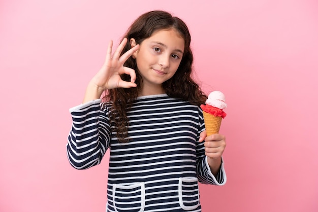 Niña caucásica sosteniendo un helado aislado en un fondo rosa mostrando el signo de ok con los dedos