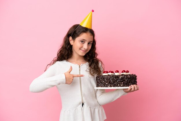 Niña caucásica sosteniendo pastel de cumpleaños aislado de fondo rosa y apuntándolo