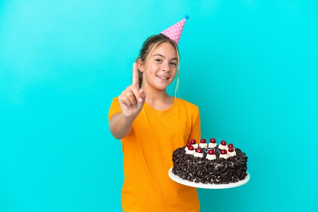 Niña caucásica sosteniendo pastel de cumpleaños aislado sobre fondo azul mostrando y levantando un dedo