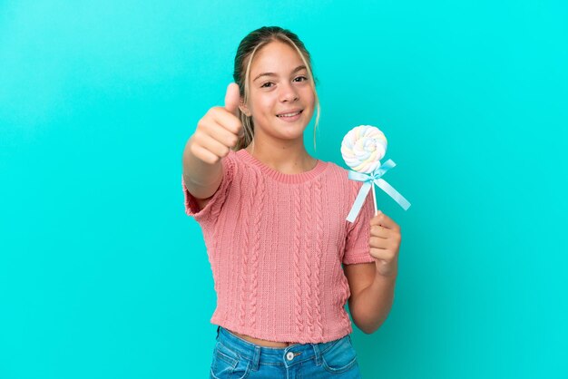 Niña caucásica sosteniendo una piruleta aislada de fondo azul con los pulgares hacia arriba porque algo bueno ha sucedido