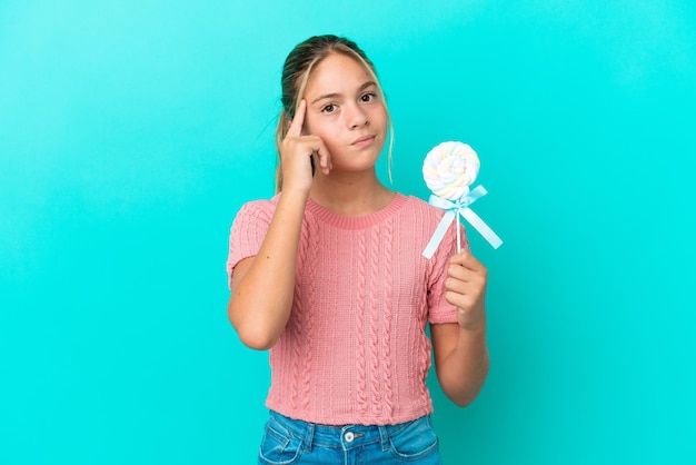 Niña caucásica sosteniendo una piruleta aislada sobre fondo azul pensando en una idea