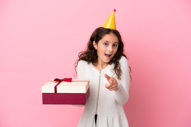 Niña caucásica sosteniendo un regalo aislado sobre fondo rosa sorprendida y apuntando al frente