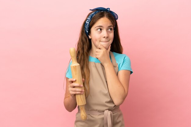 Niña caucásica sosteniendo un rodillo aislado sobre fondo rosa teniendo dudas y pensando