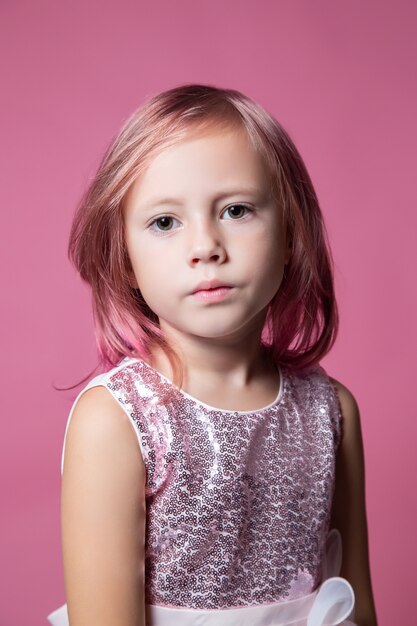 Niña caucásica en un vestido de fiesta con lentejuelas posando sobre un fondo rosa mirando a la cámara