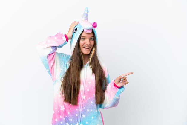 Niña caucásica vistiendo pijama de unicornio aislado sobre fondo blanco sorprendido y apuntando con el dedo hacia el lado