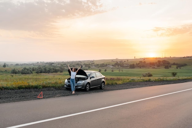 Una niña se para cerca de un auto roto en medio de la carretera durante la puesta de sol e intenta pedir ayuda por teléfono y encender el auto Esperando ayuda Servicio de auto Desglose de auto en la carretera