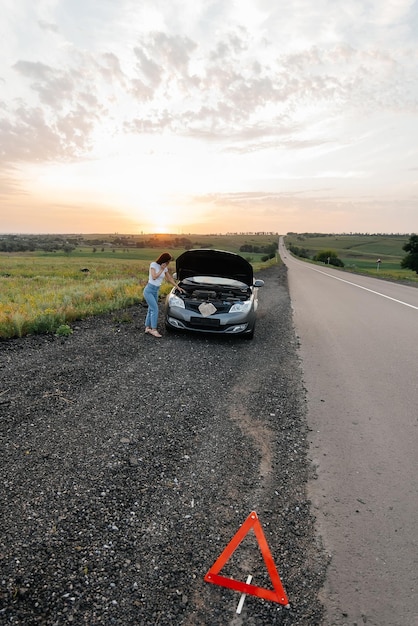 Una niña se para cerca de un auto roto en medio de la carretera durante la puesta de sol e intenta pedir ayuda por teléfono y encender el auto Esperando ayuda Servicio de auto Desglose de auto en la carretera