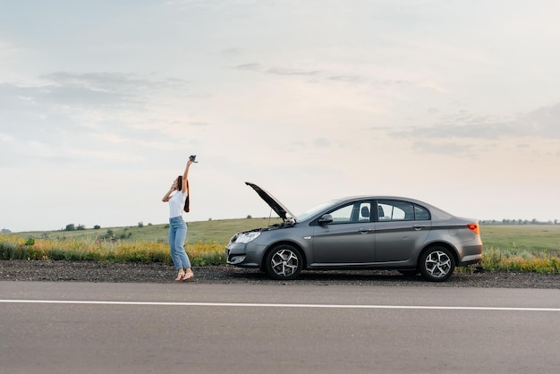 Una niña se para cerca de un auto roto en medio de la carretera durante la puesta de sol e intenta pedir ayuda por teléfono y encender el auto Esperando ayuda Servicio de auto Desglose de auto en la carretera