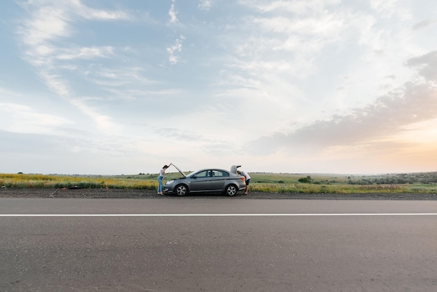 Una niña se para cerca de un automóvil averiado en medio de la carretera durante la puesta de sol e intenta pedir ayuda por teléfono Esperando ayuda Servicio de automóvil Avería de automóvil en la carretera