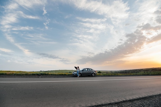 Una niña se para cerca de un automóvil averiado en medio de la carretera durante la puesta de sol e intenta pedir ayuda por teléfono Esperando ayuda Servicio de automóvil Avería de automóvil en la carretera