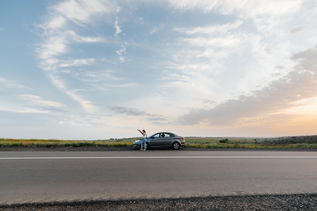 Una niña se para cerca de un automóvil averiado en medio de la carretera durante la puesta de sol e intenta pedir ayuda por teléfono Esperando ayuda Servicio de automóvil Avería de automóvil en la carretera