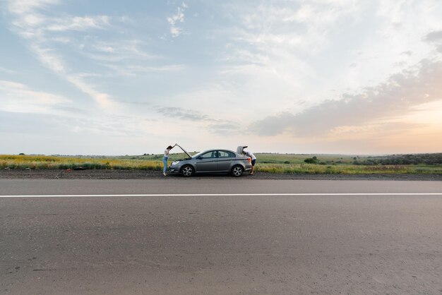 Una niña se para cerca de un automóvil averiado en medio de la carretera durante la puesta de sol e intenta pedir ayuda por teléfono Esperando ayuda Servicio de automóvil Avería de automóvil en la carretera