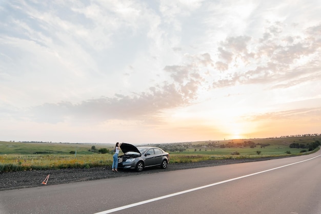 Una niña se para cerca de un automóvil averiado en medio de la carretera durante la puesta de sol e intenta pedir ayuda por teléfono Esperando ayuda Servicio de automóvil Avería de automóvil en la carretera