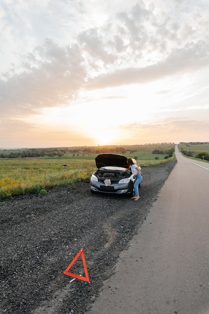 Una niña se para cerca de un automóvil roto en medio de la carretera durante la puesta de sol e intenta repararlo Solución del problema Esperando ayuda Servicio de automóvil Avería del automóvil en la carreteraxA