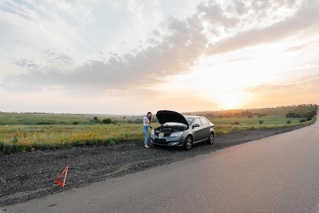 Una niña se para cerca de un automóvil roto en medio de la carretera durante la puesta de sol e intenta repararlo Solución de problemas Esperando ayuda Servicio de automóviles Avería de automóviles en la carretera