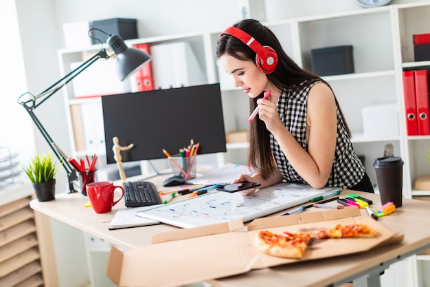 Una niña se para cerca de una mesa y tiene un marcador y un teléfono en sus manos.