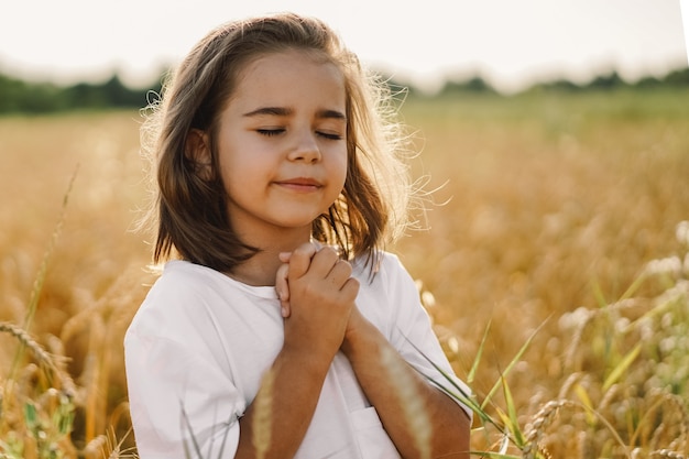 Niña cerró los ojos, rezando en un campo de trigo. Manos juntas en oración. Concepto de religión