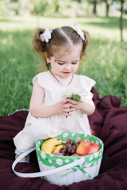 Niña con cesta de flores y frutas sentado y disfrutando día soleado de verano