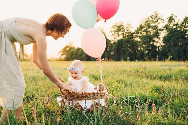 Niña en cesta de mimbre con globos a la luz del sol al atardecer Madre e hijo al aire libre Primer cumpleaños La familia celebra un bebé de un año en la naturaleza Foto de sueños saludables vacaciones vacaciones