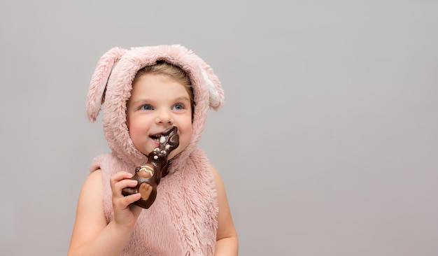 Foto niña en un chaleco con orejas de conejo come deliciosamente una liebre de chocolate en la víspera de pascua fondo gris con lugar para texto descuentos de ventas compras antes de las vacaciones