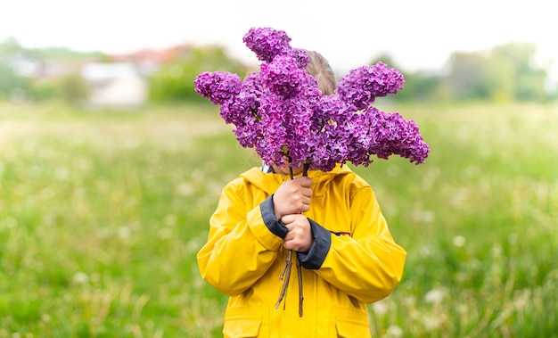 Una niña con una chaqueta amarilla se cubre la cara con un ramo de lilas.