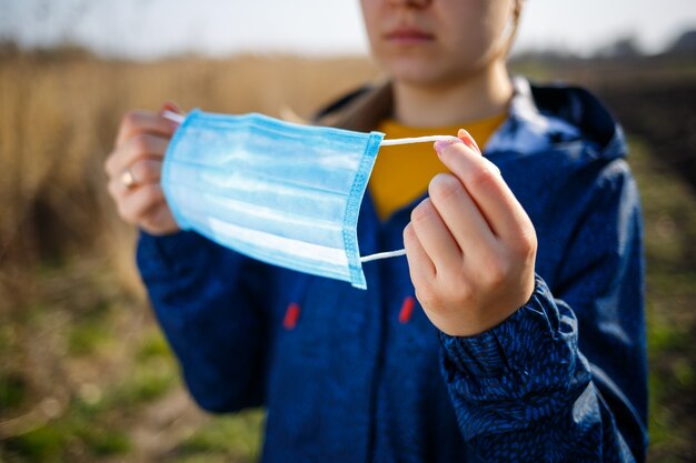 Una niña con una chaqueta azul en la naturaleza tiene una máscara protectora en sus manos. Máscara de día soleado al aire libre en la cara del virus. Enfermedad viral