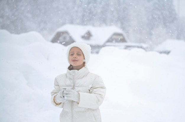 Una niña con una chaqueta blanca y un sombrero blanco sostiene una taza de té en sus manos durante una nevada en el contexto de hermosos ventisqueros y bosques