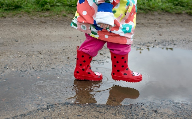 Una niña con una chaqueta brillante y botas rojas salta alegremente a través de los charcos. Humor de primavera.