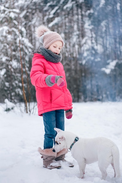 Niña con una chaqueta brillante juega en el bosque nevado de invierno con su perro jack russell terrier