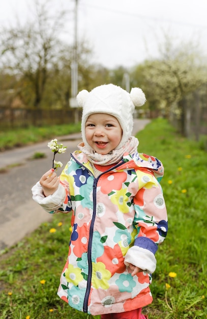 Una niña con una chaqueta brillante se ríe felizmente afuera en la primavera y sostiene una rama con flores blancas de primavera. Felicidad de los niños.