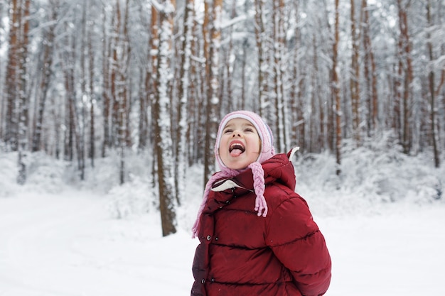Una niña con una chaqueta roja se encuentra en un bosque cubierto de nieve atrapando copos de nieve con la lengua.