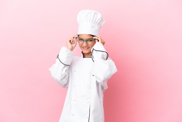 Niña chef caucásica aislada de fondo rosa con gafas y sorprendida