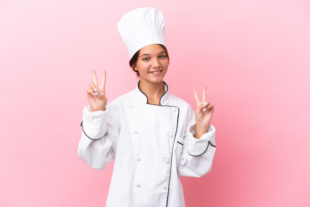 Niña chef caucásica aislada sobre fondo rosa mostrando el signo de la victoria con ambas manos