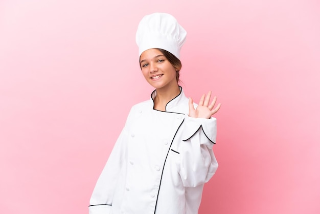 Niña chef caucásica aislada sobre fondo rosa saludando con la mano con expresión feliz
