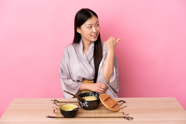 Niña china con kimono y comiendo fideos apuntando hacia el lado para presentar un producto